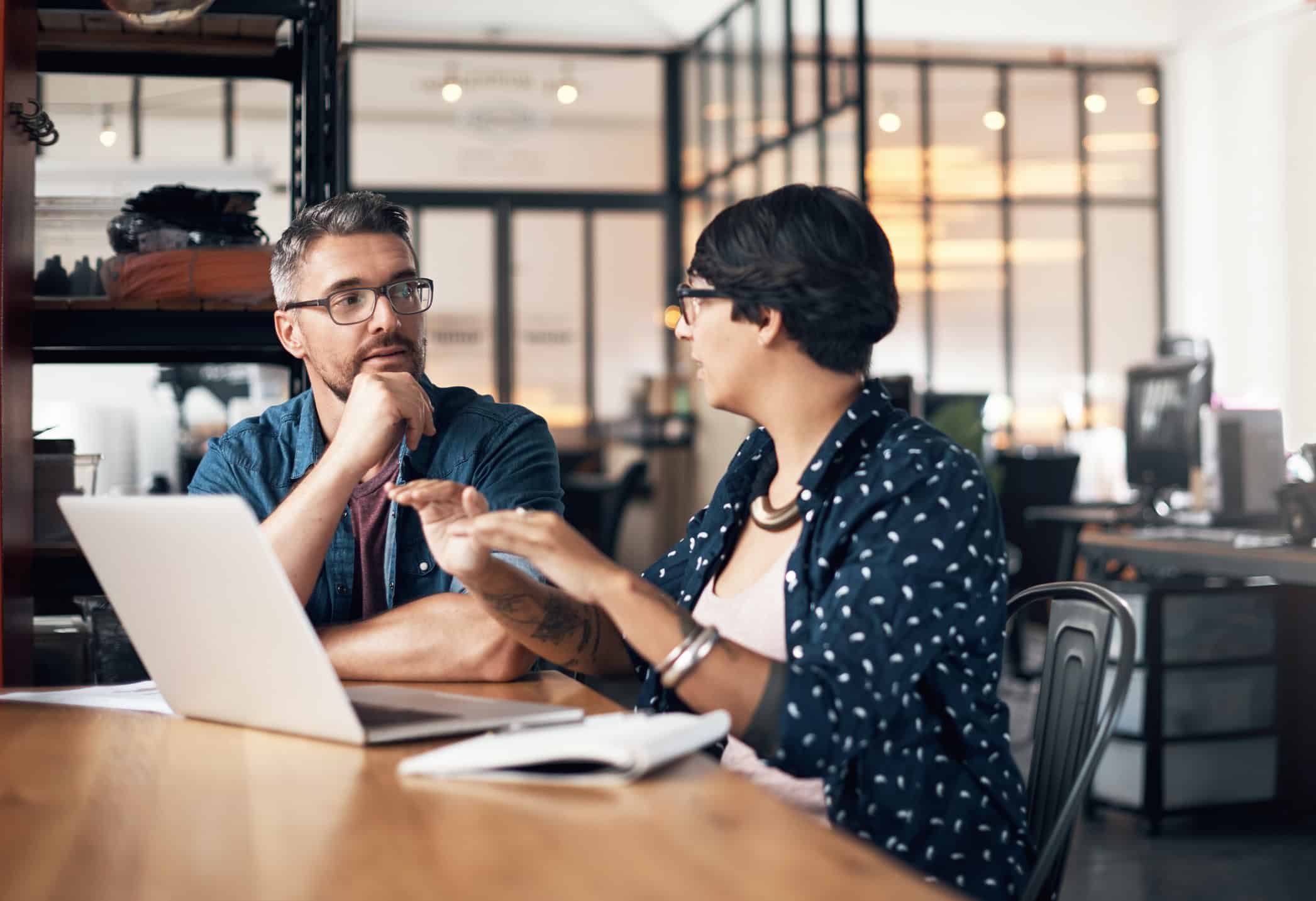 Twice the creative input on this project A man and woman seated at a desk in an office environment talking about their work