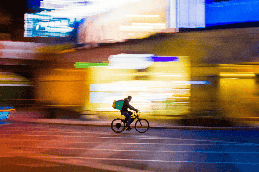 gig-economy-paolo-feser-1MFqJzNUwOM-unsplash A food delivery worker on a bicycle rides down a city road at night