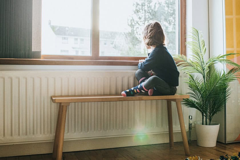 Little girl in pyjamas sitting on a bench by a sunny window inside their house. She's looking out the window