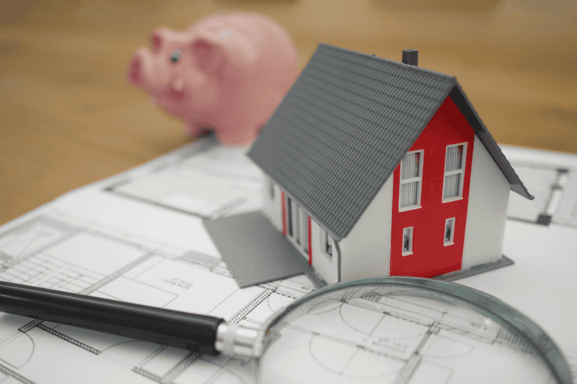 inflation-tierra-mallorca-NpTbVOkkom8-unsplash Model of a house placed on top of a housing blueprint with a piggy bank in the background