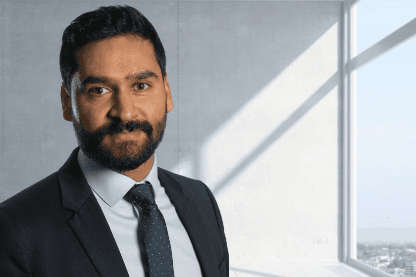 Mehul Kyprianou-Chavda wears a suit and tie. He looks relaxed, with a neutral expression. He's in a room with a concrete wall at teh back, and a large window on the right, with sunshine coming in the window.